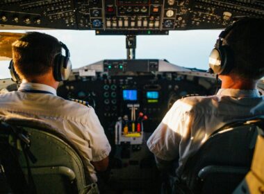 Two pilots wearing headsets operate an aircraft from the cockpit, viewed from behind