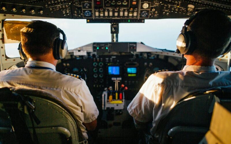 Two pilots wearing headsets operate an aircraft from the cockpit viewed from behind