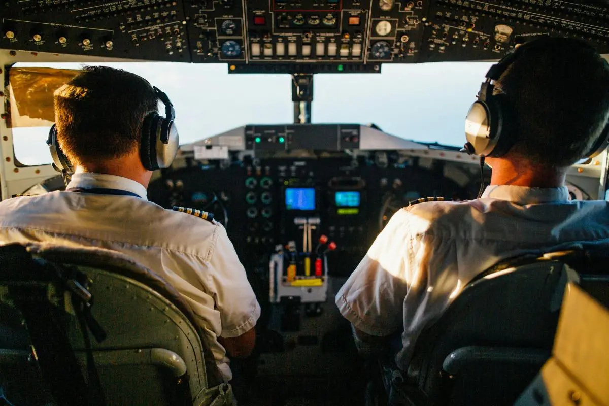 Two pilots wearing headsets operate an aircraft from the cockpit viewed from behind
