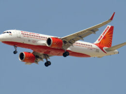 Air India passenger jet in white and orange livery taking off against a clear blue sky landing gear down and visible sunburst tail logo on the tail fin