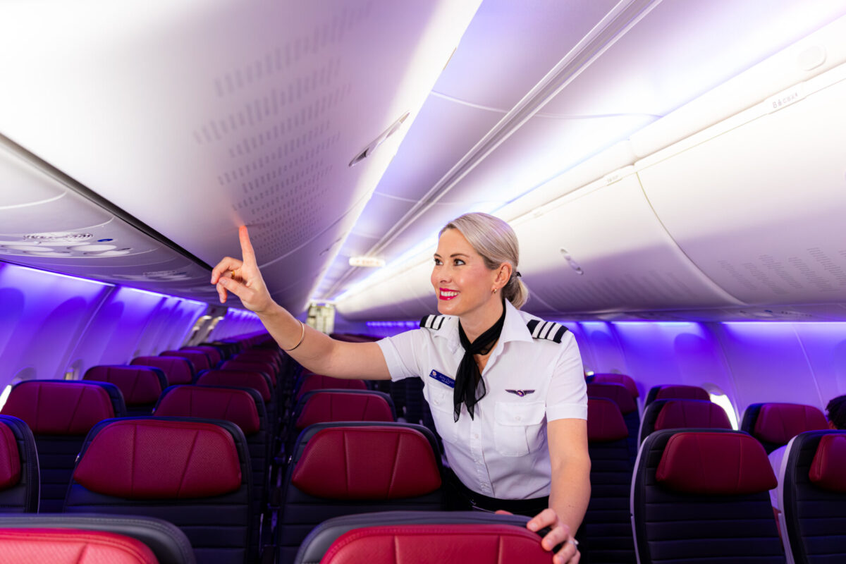 Virgin Australia 150th Boeing 737 Arrival to Australia Flight attendant with blonde hair in a white uniform points toward the overhead luggage compartment in a purple lit airplane cabin