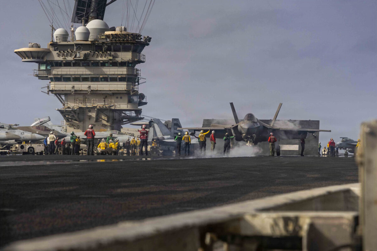 Abraham Lincoln Conducts Flight Operations Aircraft carrier flight deck with fighter jets and crew directing operations near a tall island tower under a cloudy sky with smoke at the deck edge