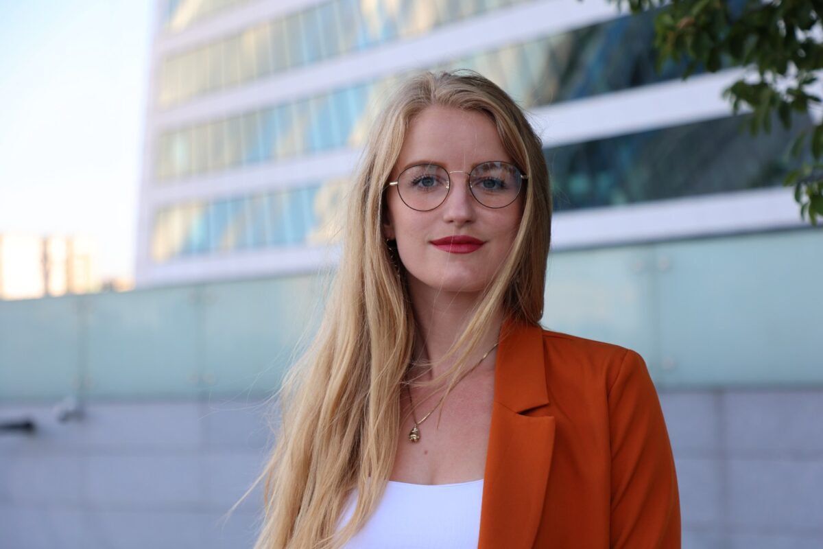 Celine Emma La Cour Woman with long blonde hair and round glasses wearing an orange blazer standing outdoors in front of a modern glass building
