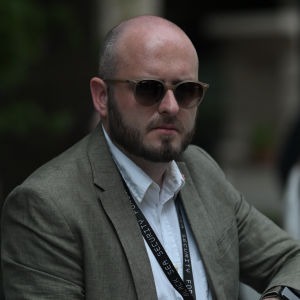 DSC 0642 imresizer Man with a shaved head and beard wearing sunglasses a light shirt and blazer a lanyard around his neck at a formal event
