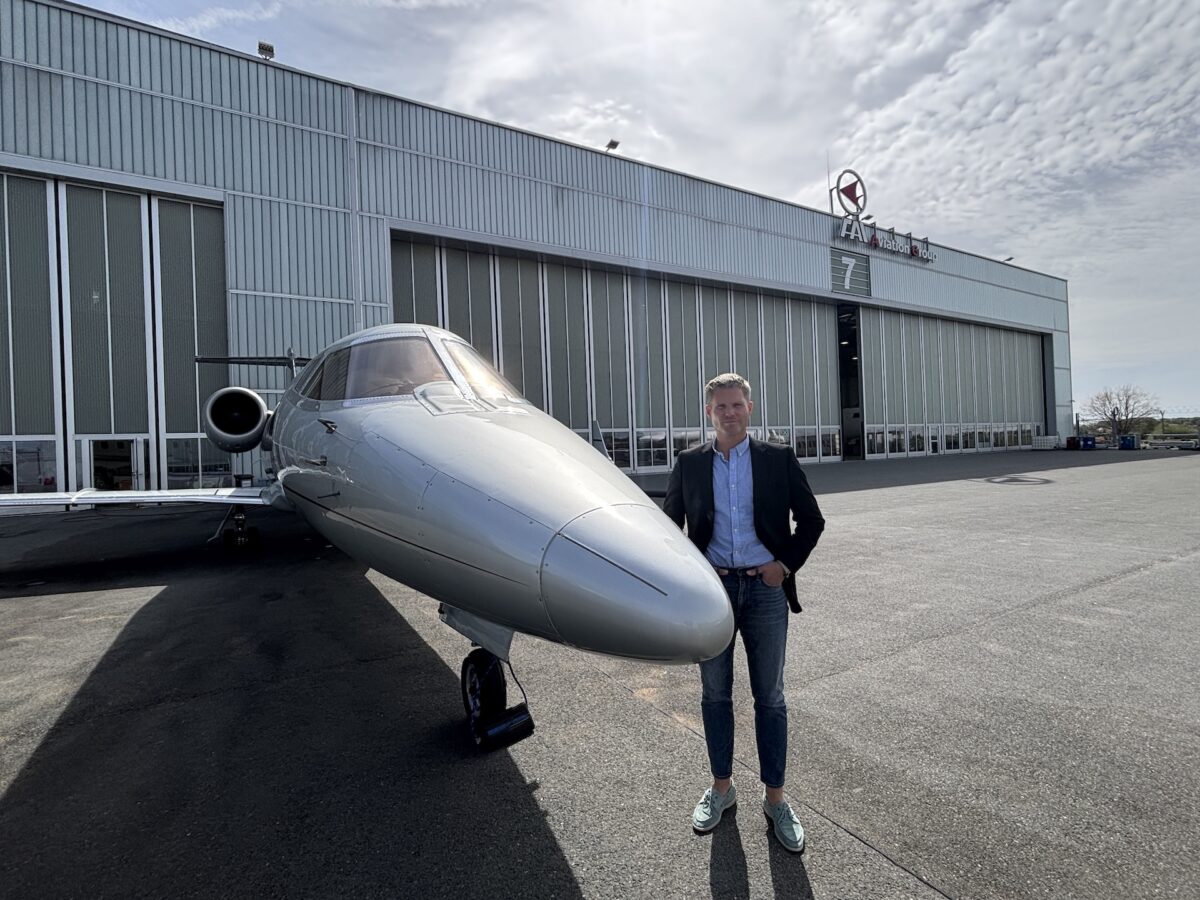 Man in a blazer posing beside a white private jet on the tarmac outside a hangar