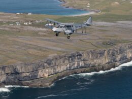 Vintage silver biplane flying along a coastal cliff with waves crashing below