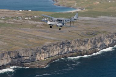 Vintage silver biplane flying along a coastal cliff with waves crashing below