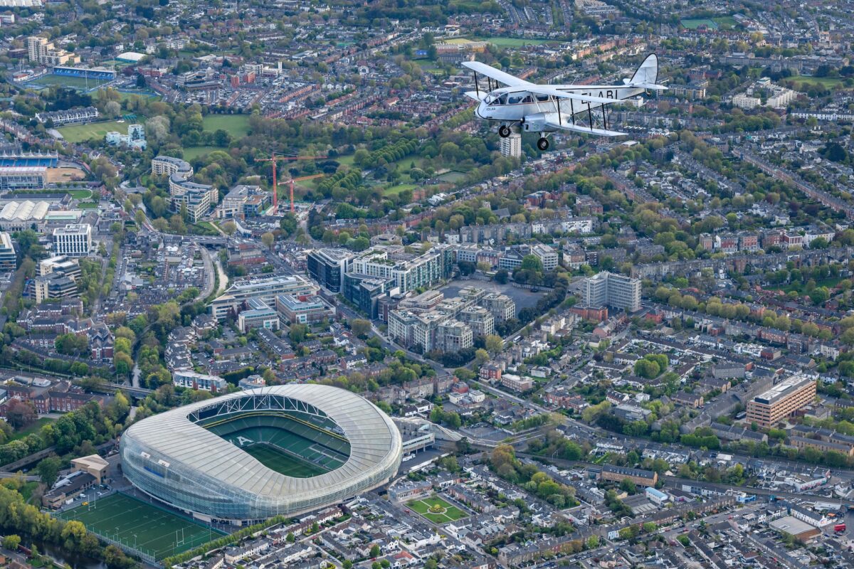A white vintage biplane flies over a dense cityscape with a large circular stadium in the foreground