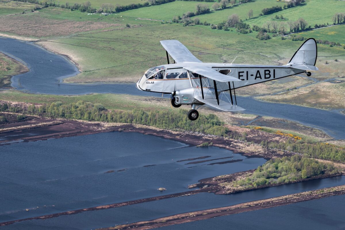 Silver vintage biplane in flight over a winding river and green landscape below registration EI ABI visible on the fuselage