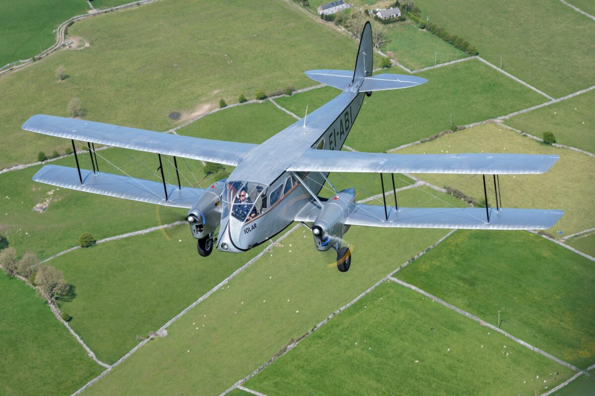 Silver twin engine biplane in flight over patchwork green fields viewed from aboveVintage gray aircraft with two wings and a cockpit flying over countrysideBiplane with registration EI ABI gliding above rural fields and stone wallsAirplane in an open sky over farmland showcasing a classic aviation silhouette