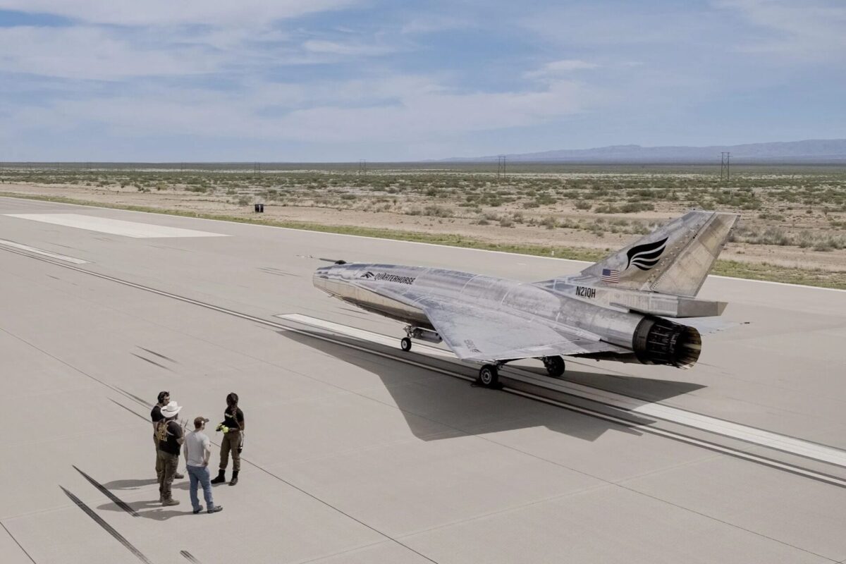 Mk21SupersonicFlightPRPhotos 9 Silver military jet parked on a wide concrete airstrip while a small group of people stands nearby with a desert landscape and distant hills in the background