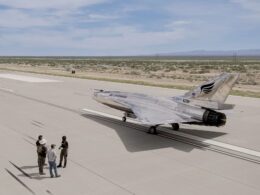 Silver military jet parked on a wide concrete airstrip while a small group of people stands nearby with a desert landscape and distant hills in the background
