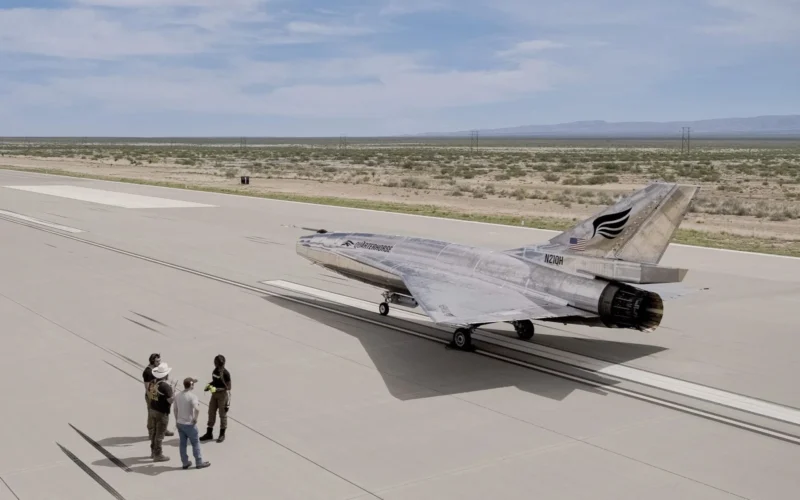 Silver military jet parked on a wide concrete airstrip while a small group of people stands nearby with a desert landscape and distant hills in the background