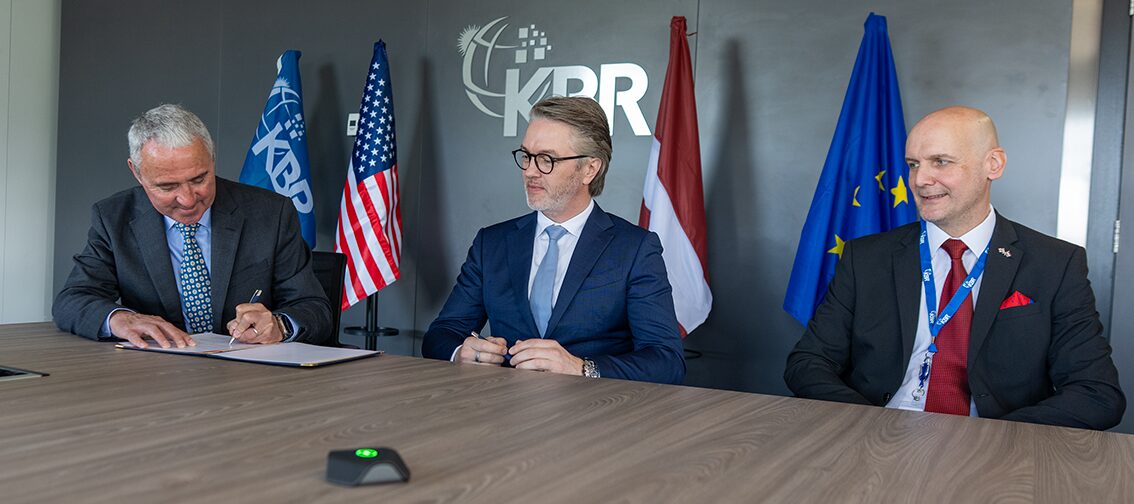 NorSAF and KBR Sign Agreement Three men in suits sit at a conference table the left man signs a document while the others watch with flags and a logo backdrop behind them