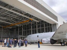 Group of people standing on the tarmac near a gray jet inside a large hangar with a yellow overhead crane above