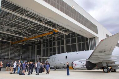 Group of people standing on the tarmac near a gray jet inside a large hangar with a yellow overhead crane above