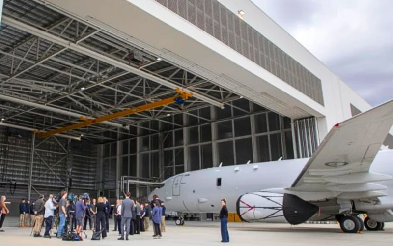 Group of people standing on the tarmac near a gray jet inside a large hangar with a yellow overhead crane above