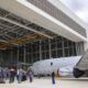 Group of people standing on the tarmac near a gray jet inside a large hangar with a yellow overhead crane above