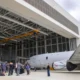 Group of people standing on the tarmac near a gray jet inside a large hangar with a yellow overhead crane above