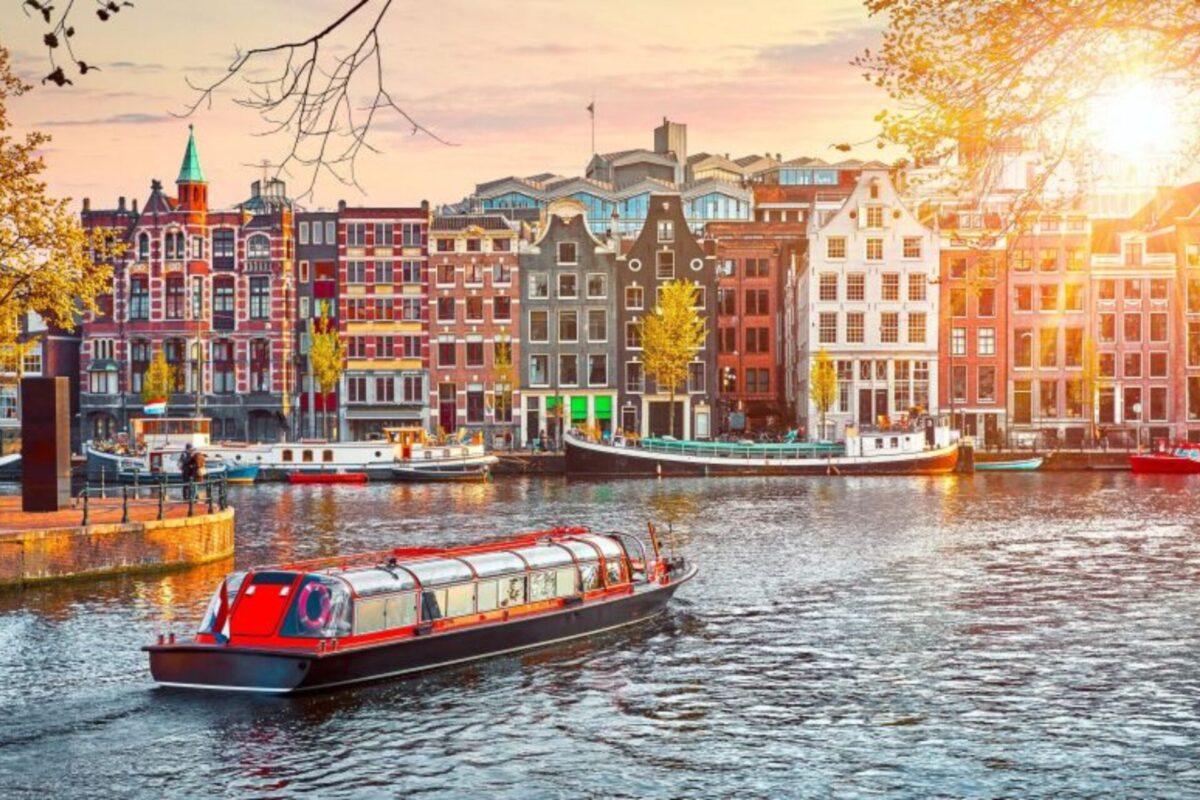 Canal in Amsterdam with colorful row of historic narrow houses and a red roofed boat cruising the water at sunset