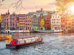 Canal in Amsterdam with colorful row of historic narrow houses and a red roofed boat cruising the water at sunset