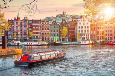 Canal in Amsterdam with colorful row of historic narrow houses and a red roofed boat cruising the water at sunset
