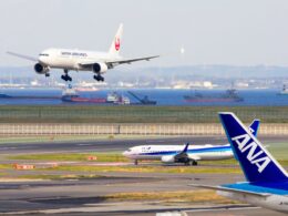 Japan Airlines jet landing over a harbor with ships on the water and an ANA tail in the foreground