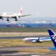 Japan Airlines jet landing over a harbor with ships on the water and an ANA tail in the foreground