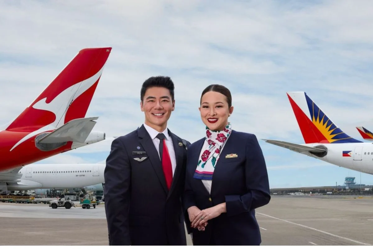 Two airline crew members a man and a woman in dark uniforms stand smiling on a runway with red and white aircraft tails in the background