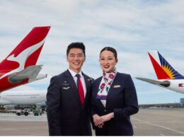 Two airline crew members a man and a woman in dark uniforms stand smiling on a runway with red and white aircraft tails in the background