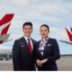 Two airline crew members a man and a woman in dark uniforms stand smiling on a runway with red and white aircraft tails in the background