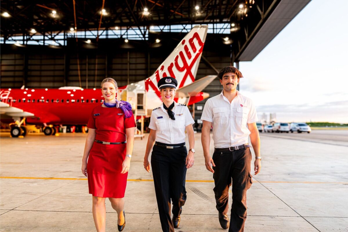 Three airline staff walking toward the camera in a hangar with a red aircraft in the background