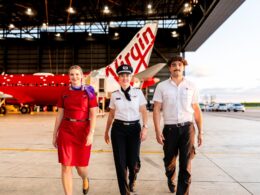 Three airline staff walking toward the camera in a hangar with a red aircraft in the background