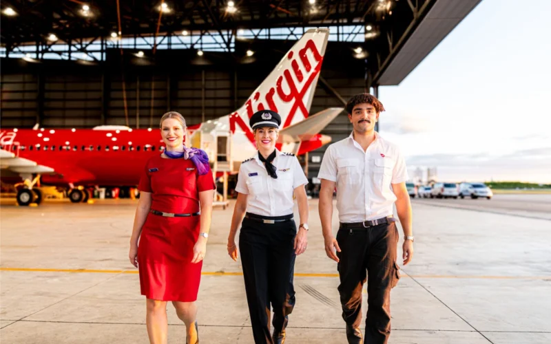 Three airline staff walking toward the camera in a hangar with a red aircraft in the background