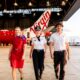 Three airline staff walking toward the camera in a hangar with a red aircraft in the background