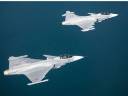 Three gray fighter jets fly in formation over a calm blue ocean viewed from above with crews visible in the cockpits