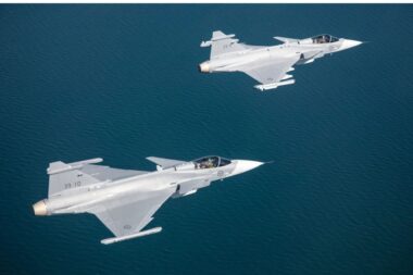Three gray fighter jets fly in formation over a calm blue ocean viewed from above with crews visible in the cockpits