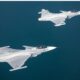 Three gray fighter jets fly in formation over a calm blue ocean viewed from above with crews visible in the cockpits