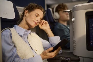 Woman asleep with hand to temple holding a tablet wearing a light quilted vest over a blue shirt on an airplane seat