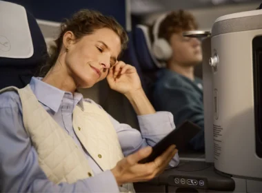 Woman asleep with hand to temple holding a tablet wearing a light quilted vest over a blue shirt on an airplane seat