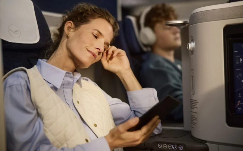 Woman asleep with hand to temple holding a tablet wearing a light quilted vest over a blue shirt on an airplane seat