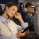 Woman asleep with hand to temple holding a tablet wearing a light quilted vest over a blue shirt on an airplane seat