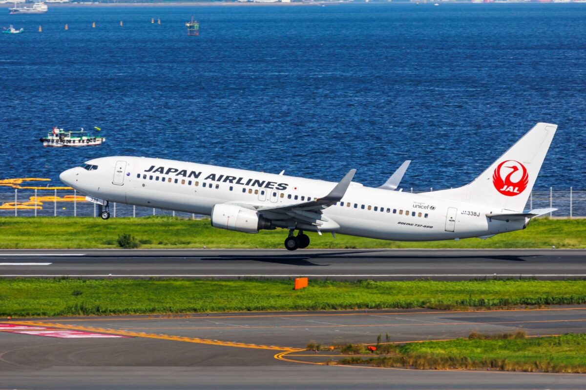 Japan Airlines Boeing 737 800 taking off on a runway beside a blue harbor with the JAL red logo on the tail and green grass nearby