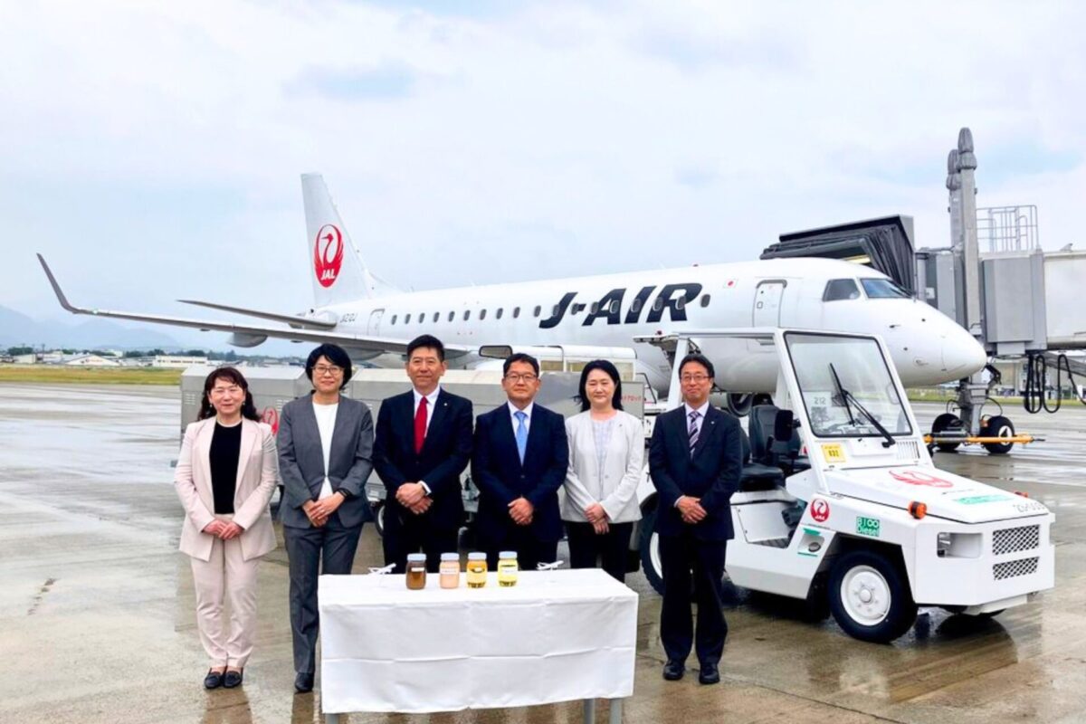 Seven professionals in business attire stand in a line in front of a Japan Airlines jet at the gate with a table of bottled drinks in front