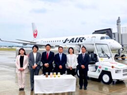 Seven professionals in business attire stand in a line in front of a Japan Airlines jet at the gate with a table of bottled drinks in front