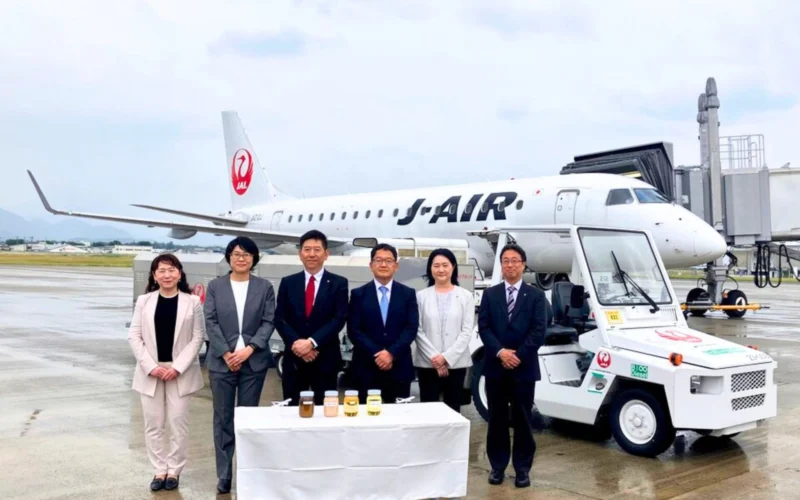 Seven professionals in business attire stand in a line in front of a Japan Airlines jet at the gate with a table of bottled drinks in front