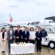 Seven professionals in business attire stand in a line in front of a Japan Airlines jet at the gate with a table of bottled drinks in front