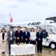 Seven professionals in business attire stand in a line in front of a Japan Airlines jet at the gate with a table of bottled drinks in front