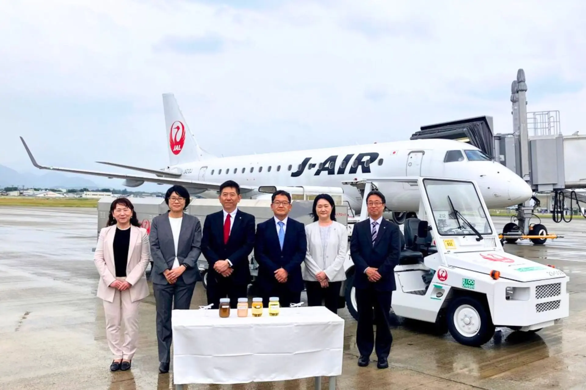 Seven professionals in business attire stand in a line in front of a Japan Airlines jet at the gate with a table of bottled drinks in front