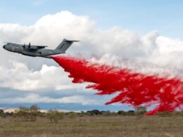 Military transport airplane releasing bright red smoke trail over an open field